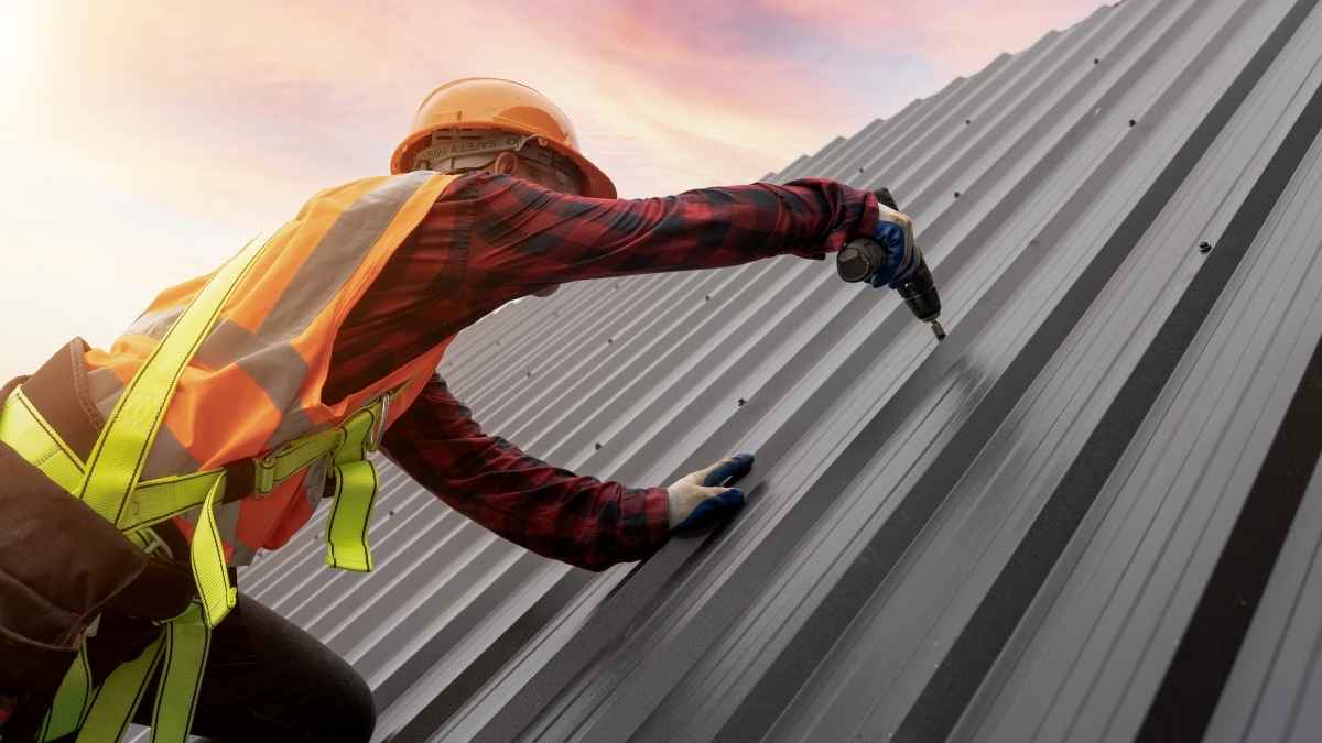 A construction worker in a safety vest and helmet uses a power drill to install or repair a metal roof at sunset.