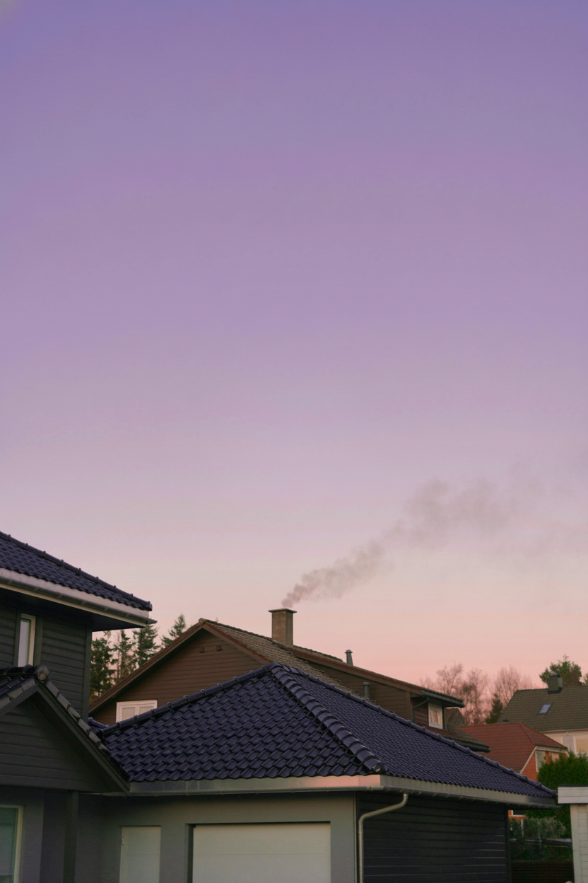 A house with smoke rising from the chimney sits under a purple and pink sky at sunset. Other rooftops and trees are partially visible in the background.