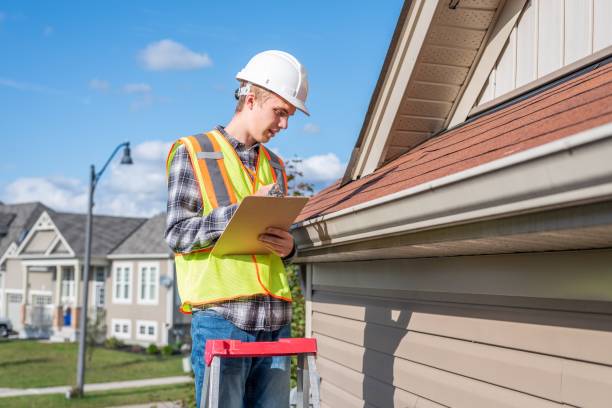 A construction worker wearing a hard hat and safety vest stands on a ladder, inspecting the roof and gutter of a house while writing on a clipboard in a suburban neighborhood.
