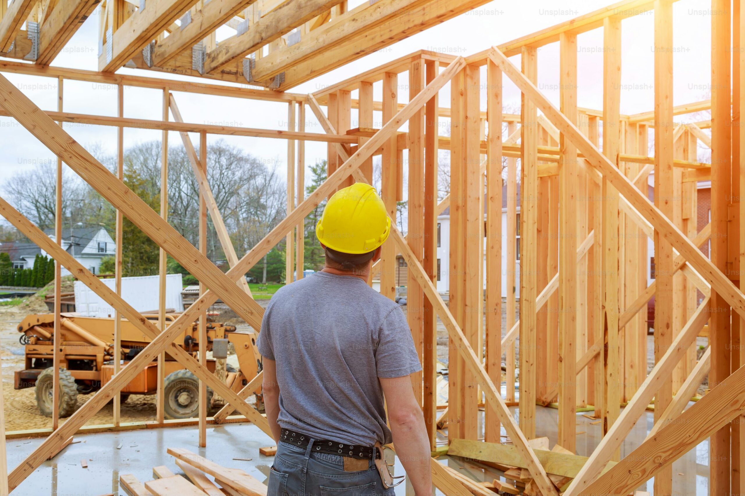 A construction worker in a yellow hard hat and gray shirt stands inside a wooden frame of a house under construction, looking up at the structure on a sunny day.