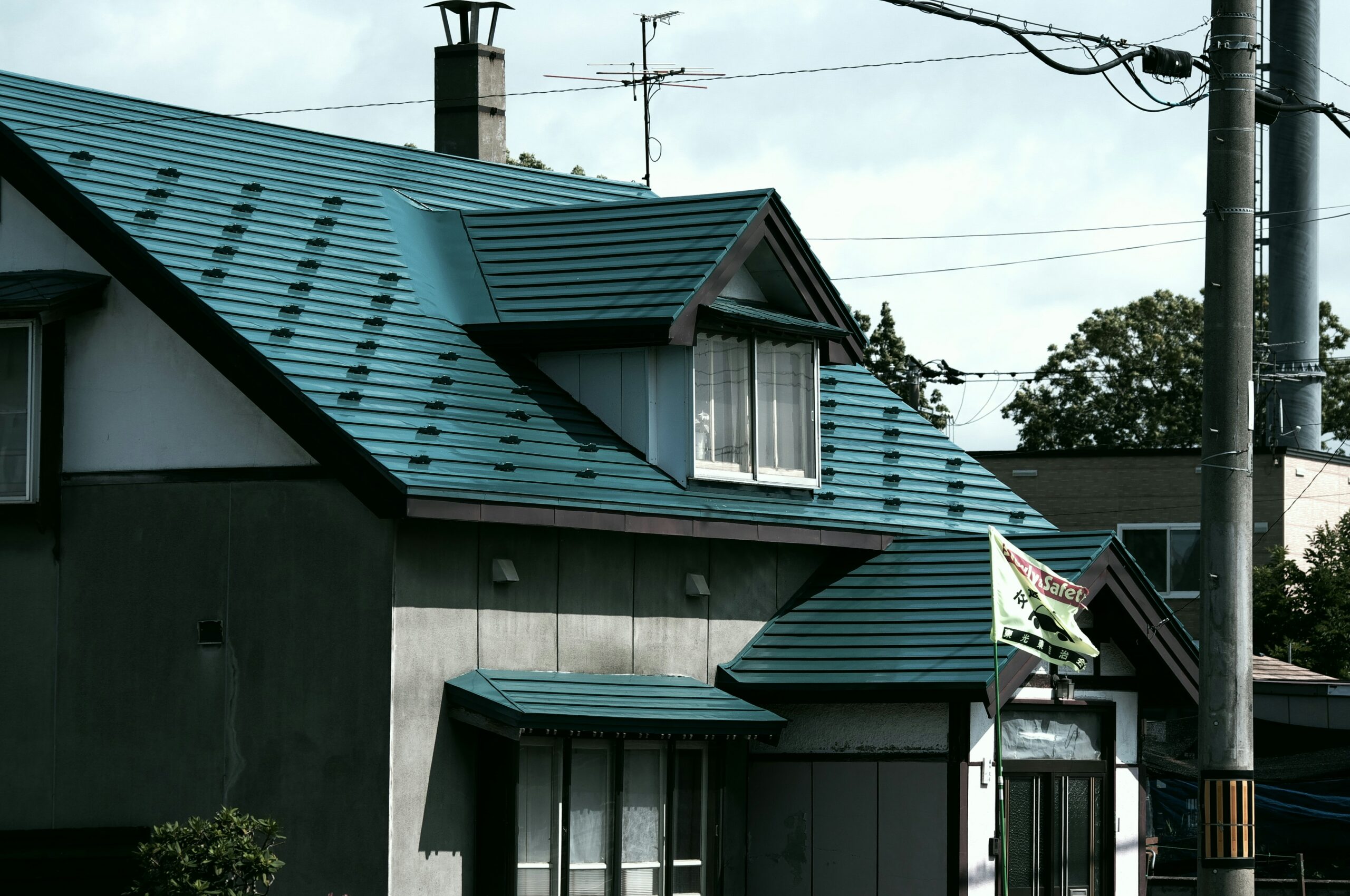 A house with teal roofing and dormer windows, featuring an outdoor light fixture, power lines, and two green flags hanging by the entrance. Trees and another building are visible in the background.