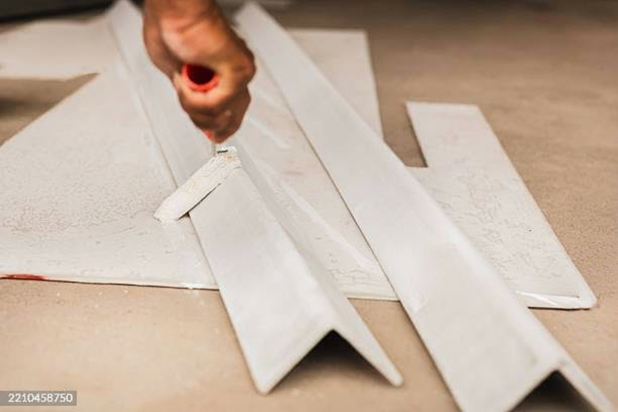 A hand using a roller brush to paint long, white metal or wooden angle strips placed on a flat surface, with some paint visible on the roller and pieces.
