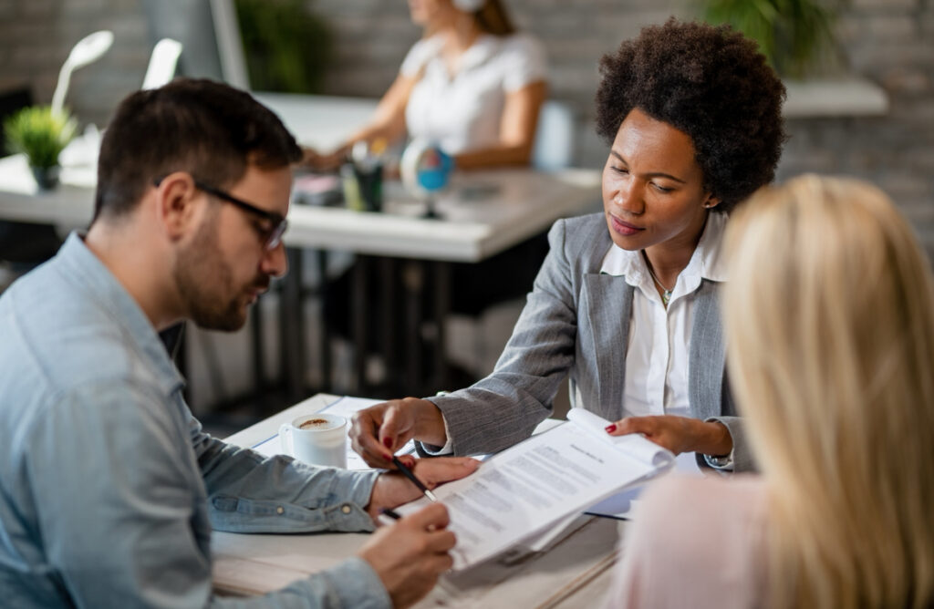 A businesswoman in a gray blazer explains documents to a man and woman at a desk in a modern office, with another person working in the background.