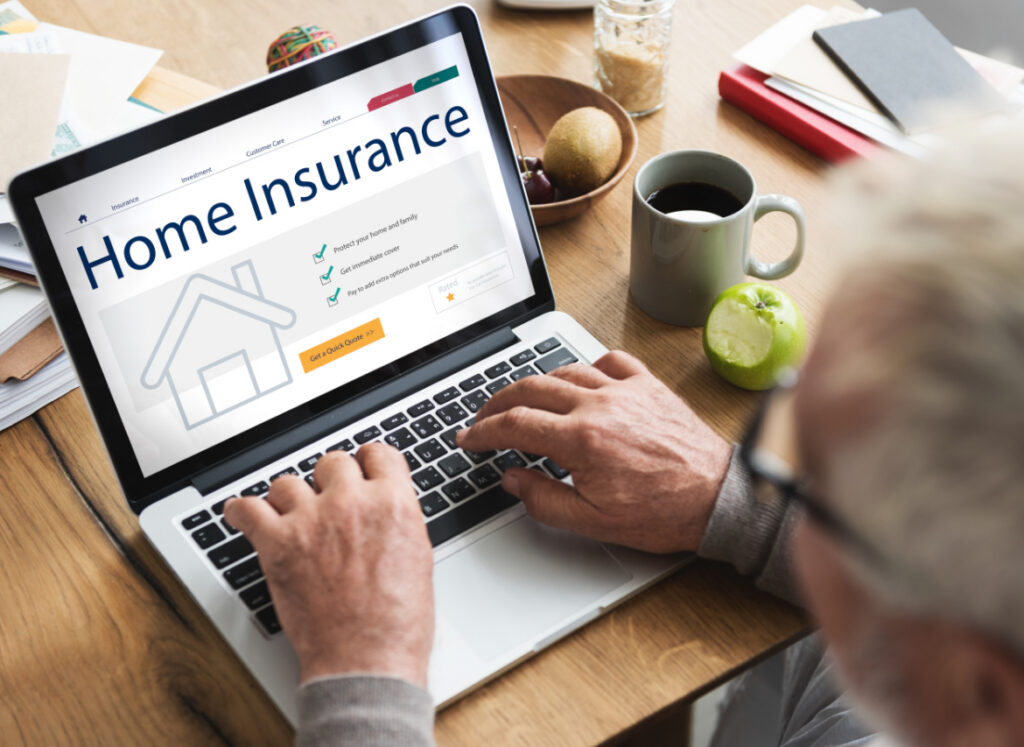 An older person uses a laptop displaying a home insurance website. The screen shows checkmarks next to covered items, with a coffee mug, green apple, glasses, and papers on the wooden table nearby.