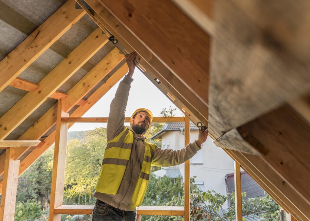 A construction worker in a yellow vest and hard hat uses a level tool to check the alignment of a wooden roof frame on a building under construction. Trees and a house are visible in the background.