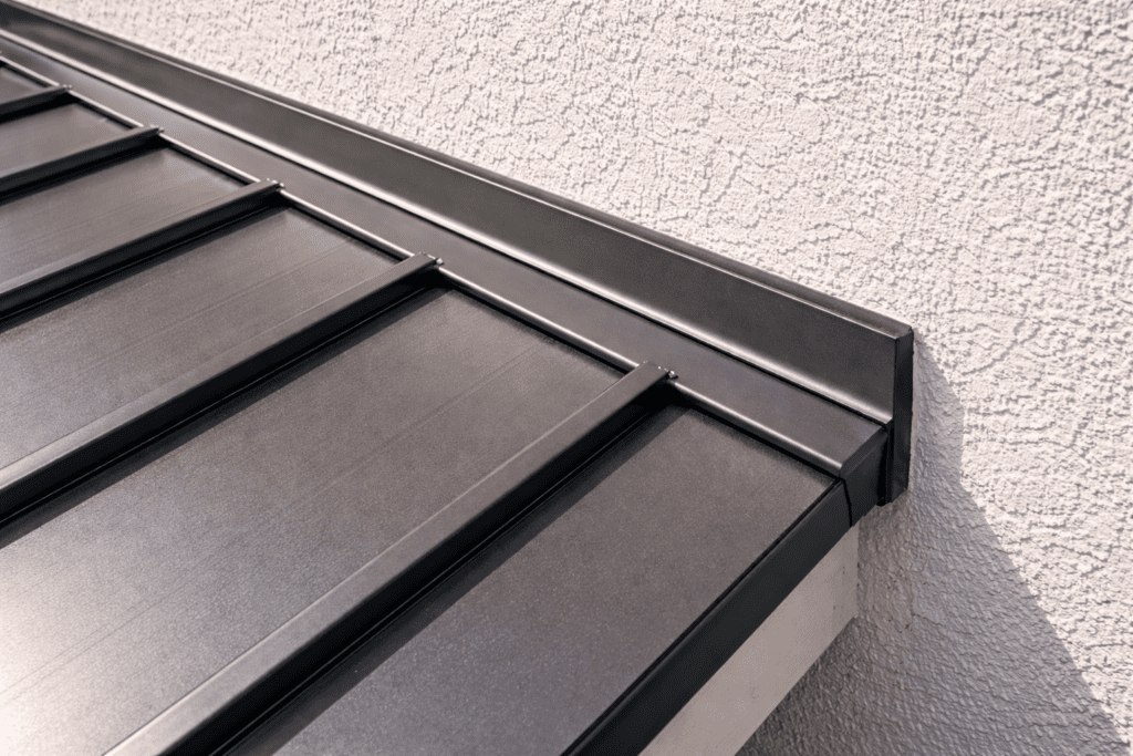 Close-up of a modern black metal roof with standing seam panels, attached to a light-colored textured exterior wall of a building. The image highlights the clean lines and precise installation of the roofing.