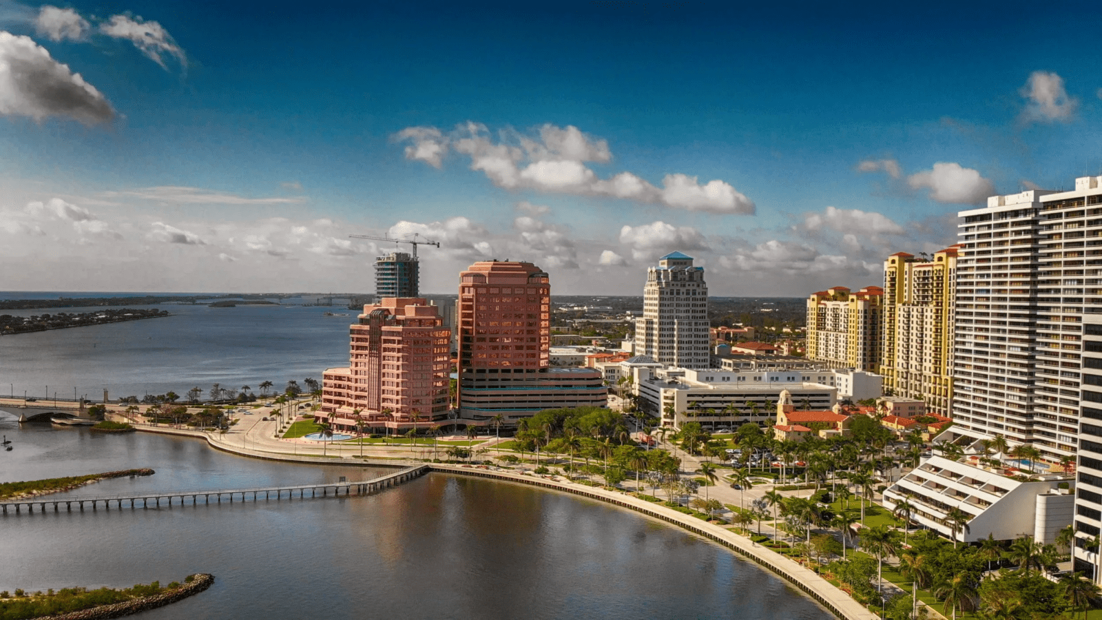 Aerial view of a waterfront cityscape with tall buildings, palm trees, and a curved walkway along the shoreline under a partly cloudy blue sky.