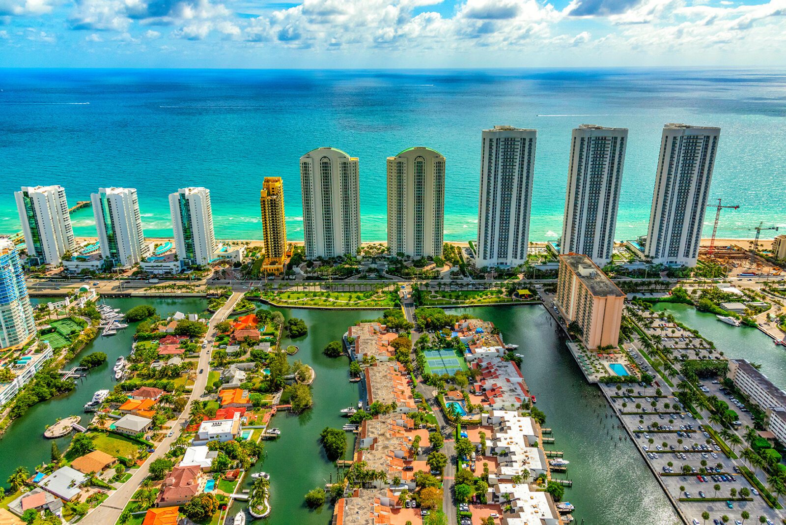 Aerial view of a coastal city with tall modern skyscrapers along the beach, turquoise ocean, waterways, and residential homes with boats docked nearby under a partly cloudy sky.