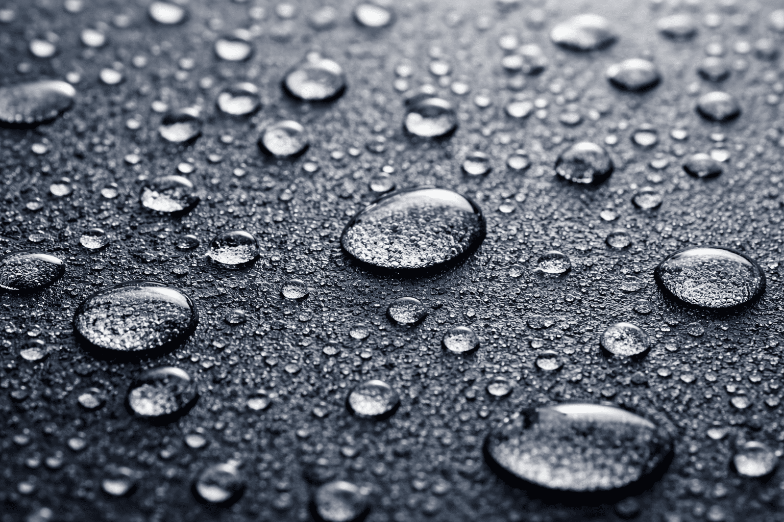 Close-up of water droplets of varying sizes scattered on a dark, smooth surface, creating a reflective and textured appearance. The surface and droplets catch the light, highlighting their shiny and glossy texture.