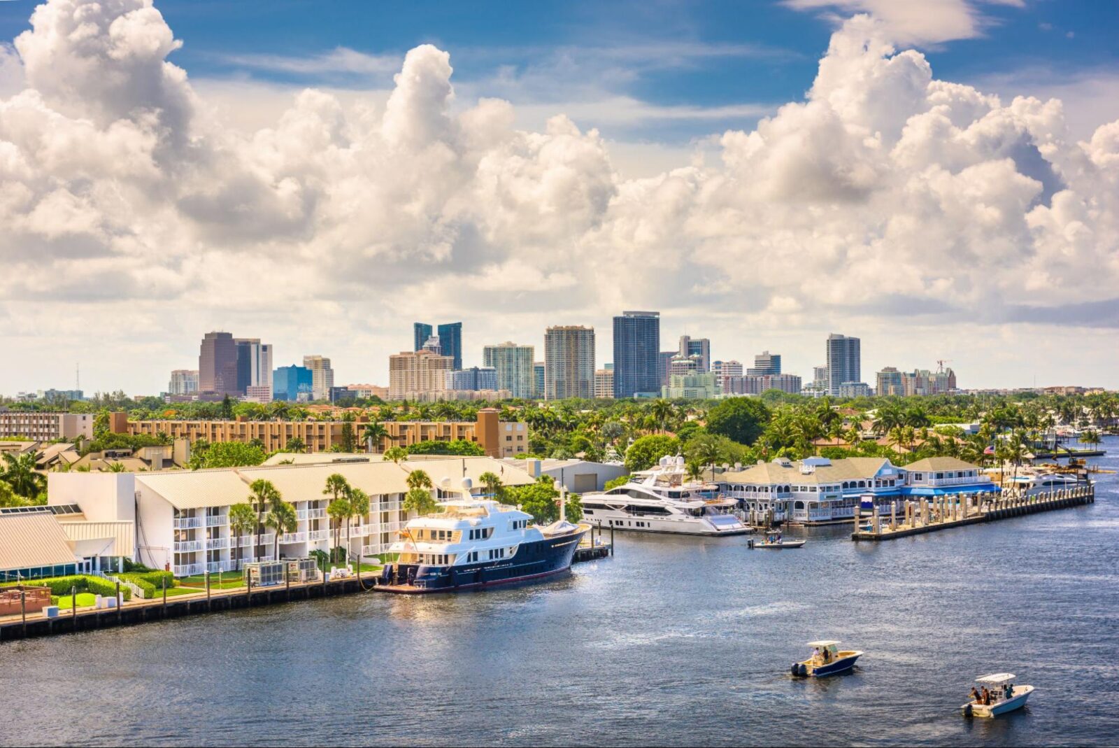 A waterfront cityscape with boats and yachts docked along a marina, palm trees, and a skyline of modern high-rise buildings under a partly cloudy sky.