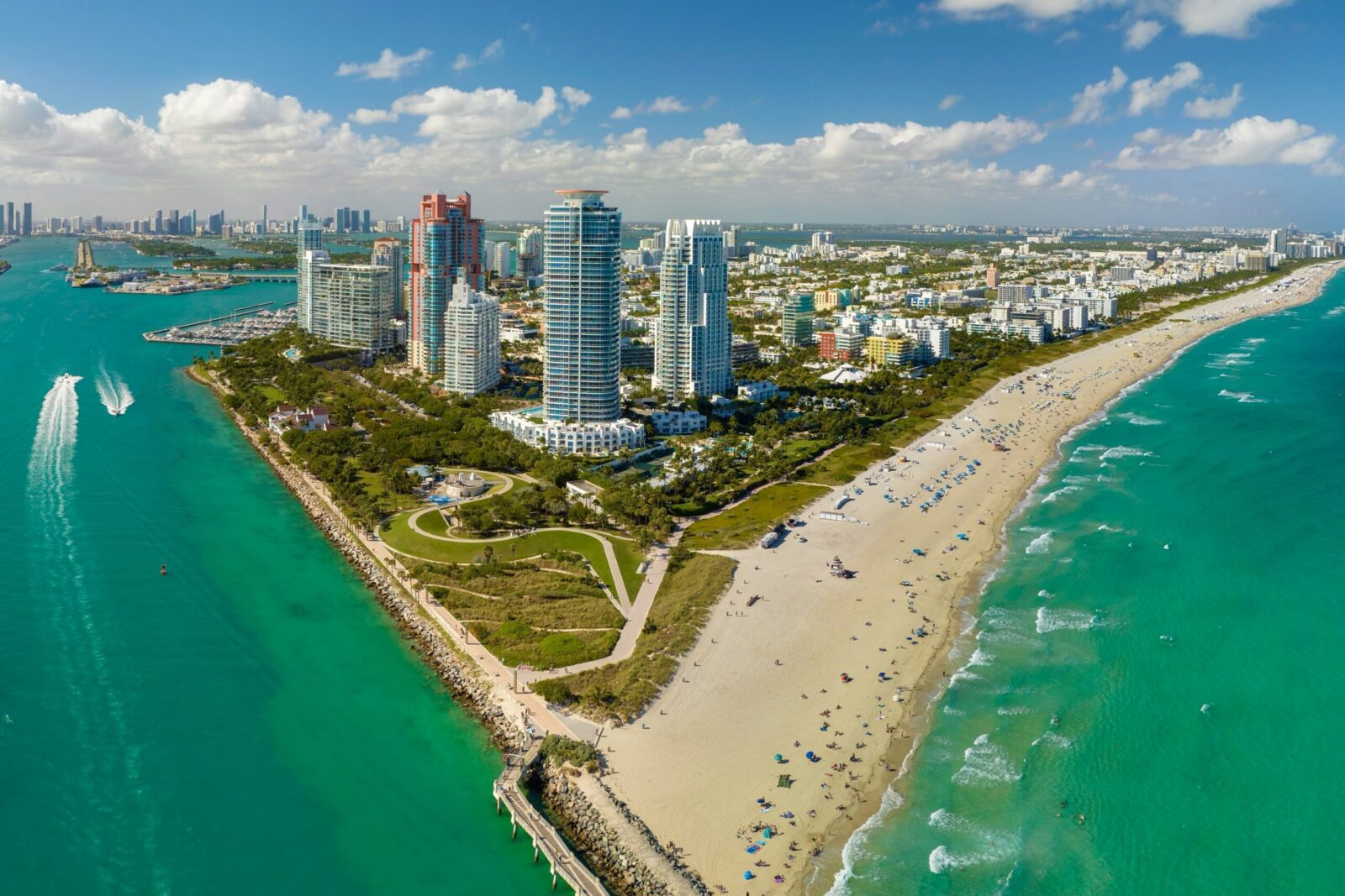 Aerial view of a city skyline with tall buildings along a sandy beach, bordered by turquoise ocean water. People are on the beach and boats are visible in the water. Green park areas separate the beach from the buildings.