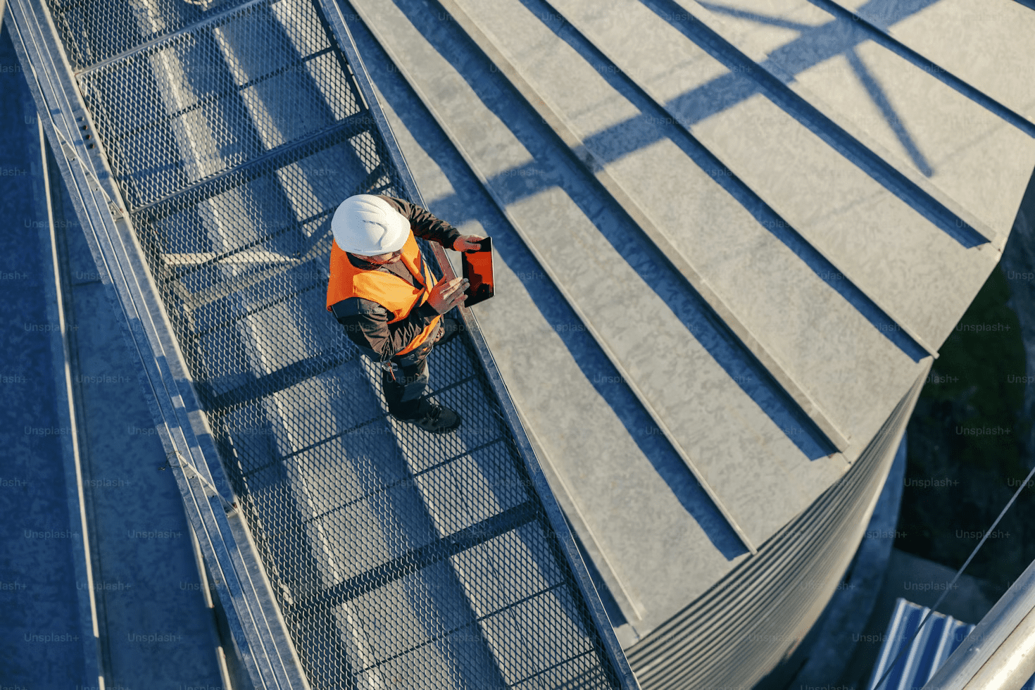 A construction worker in a white hard hat and orange safety vest stands on a metal walkway, holding a red clipboard near a large industrial structure with metal panels.