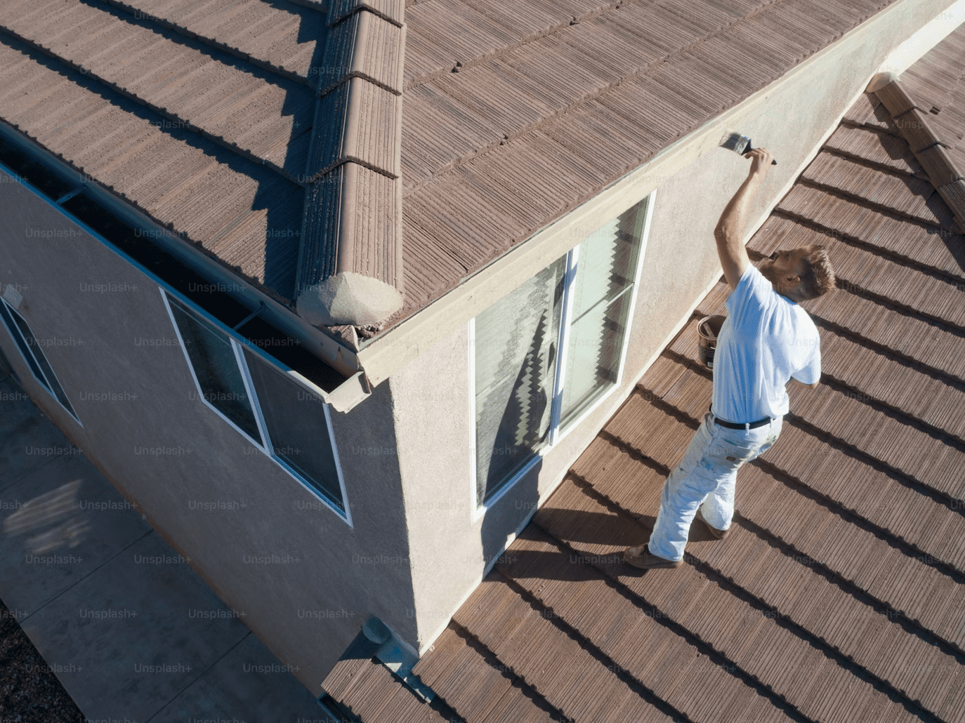 A person dressed in white is standing on the roof of a house, painting the trim near the roofline with a brush. The house has brown roof tiles and light-colored exterior walls.