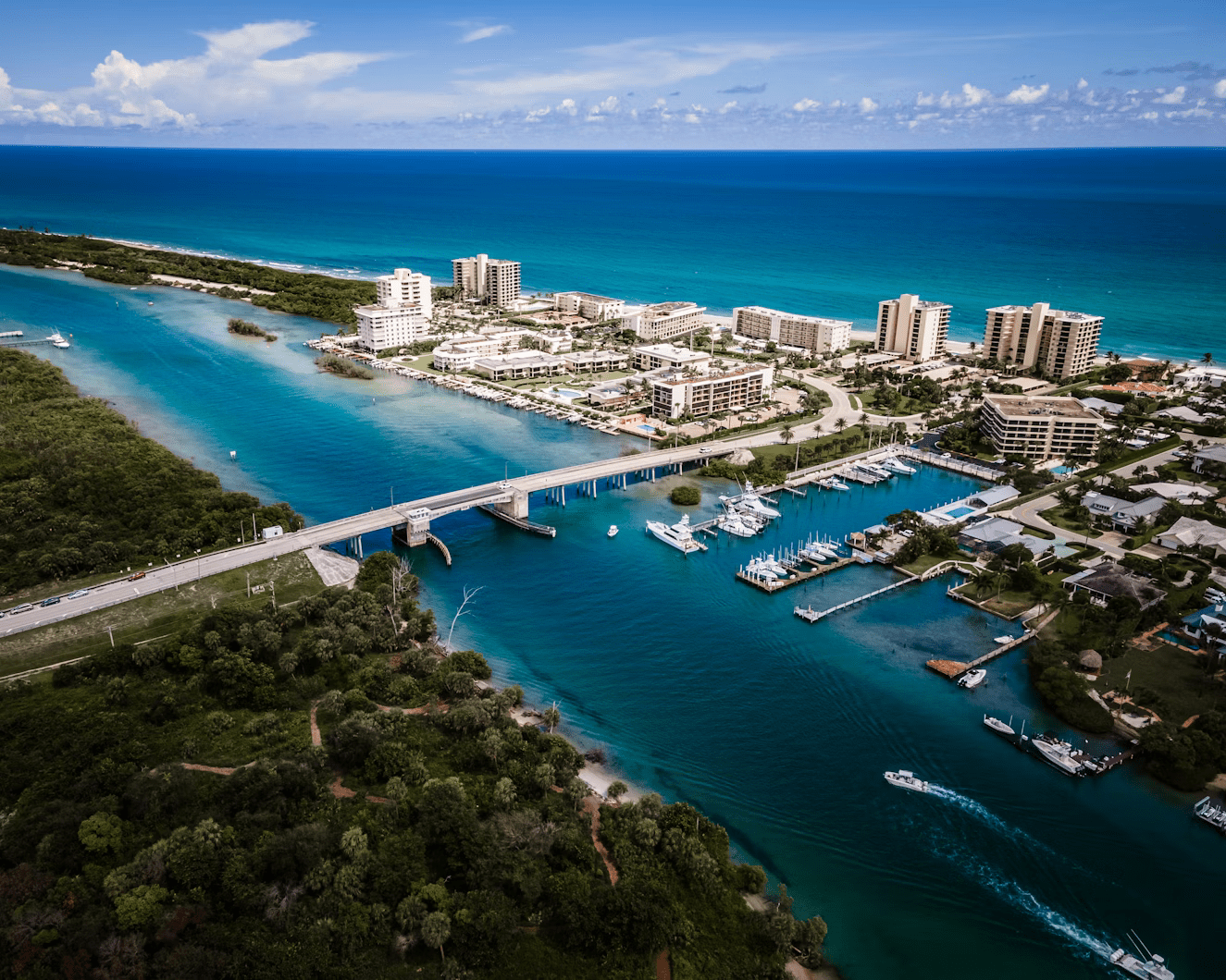 Aerial view of a coastal city with a bridge spanning blue-green water, connecting two land areas. High-rise buildings and a marina with boats are visible along the shoreline, with a lush, green area in the foreground.