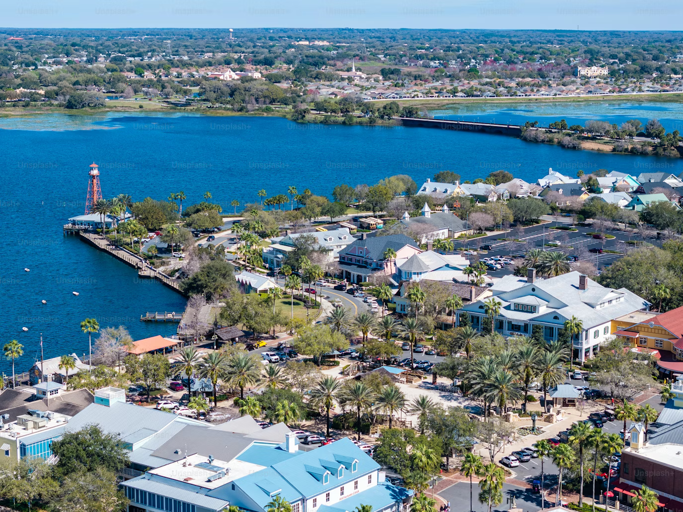Aerial view of a waterfront town with colorful buildings, palm trees, parked cars, and a small red lighthouse on a point by a lake, with a bridge and more greenery in the background.