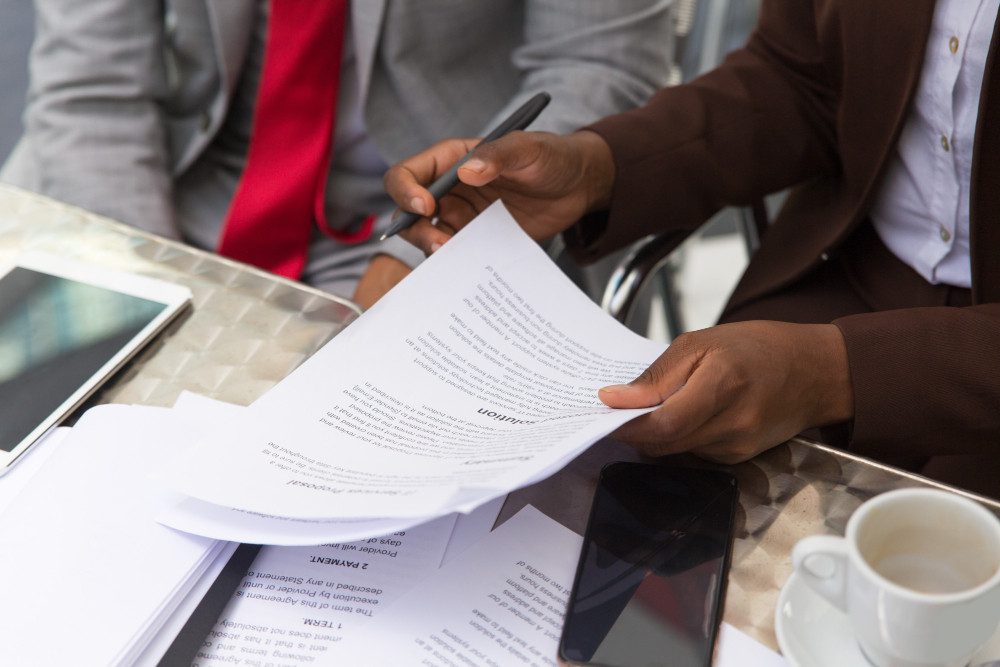 Two people in business attire review documents at a table. One holds papers and a pen, while a phone, tablet, and cup of coffee are nearby. Only their hands and part of their torsos are visible.
