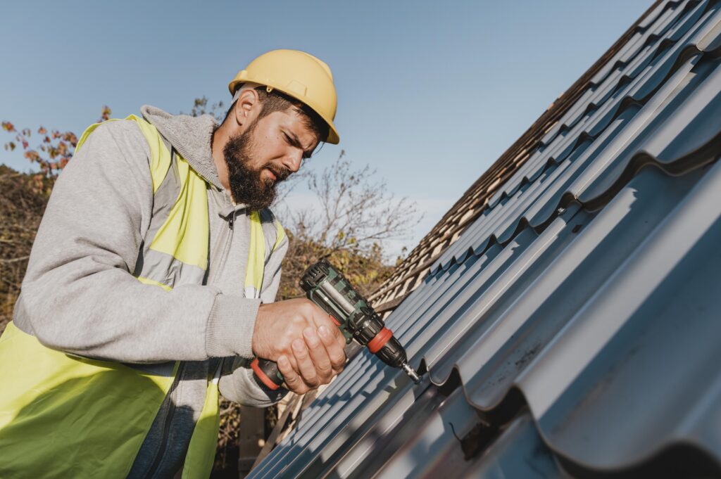 A construction worker wearing a yellow hard hat and reflective vest uses a cordless drill to fasten metal roofing sheets on a sloped roof under a clear blue sky.