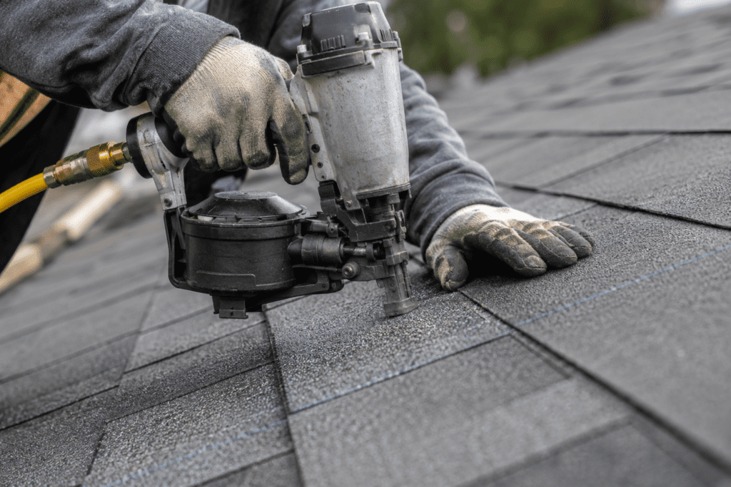 A person wearing work gloves uses a pneumatic nail gun to install asphalt shingles on a roof, with rows of shingles visible and a blurred background of greenery.