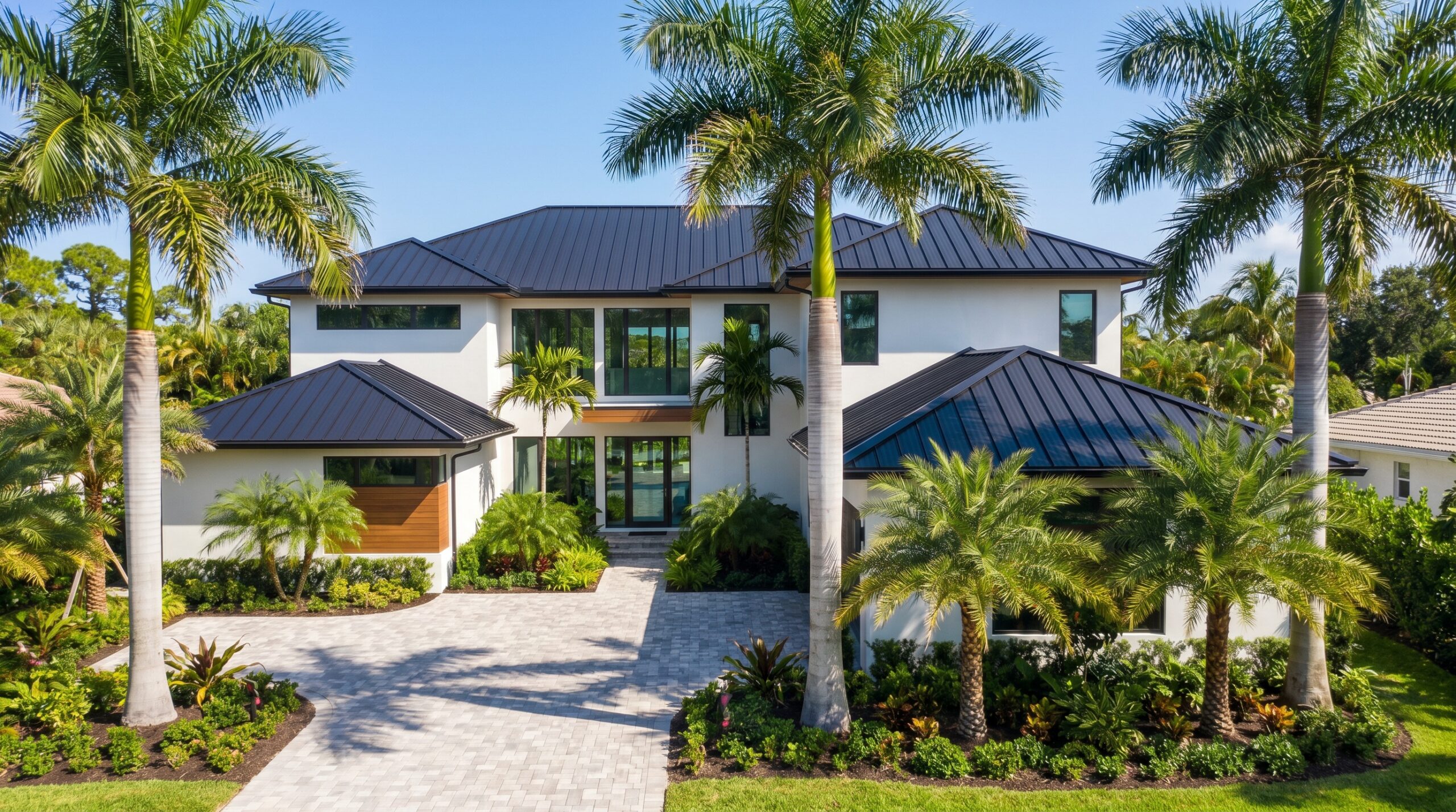 A modern two-story house with a dark metal roof, large windows, and white exterior, surrounded by lush landscaping and tall palm trees, with a paved driveway leading to the entrance.