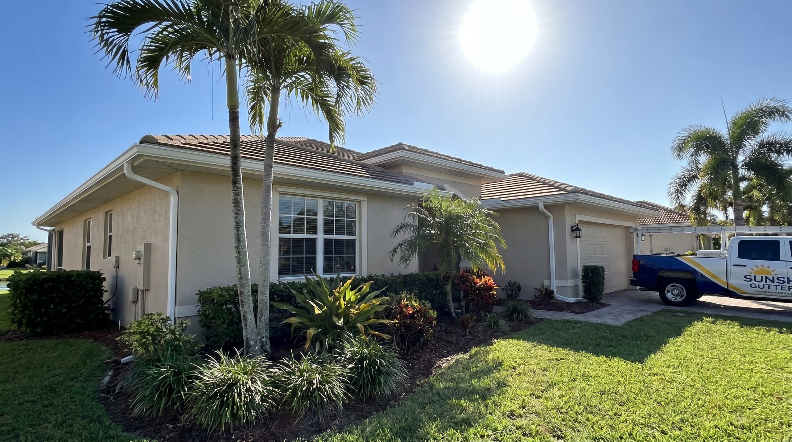 A beige single-story house with a tile roof, surrounded by green lawn, palm trees, and shrubs under a bright, sunny sky. A blue and yellow truck with “Sunshine Gutters” is parked in the driveway.
