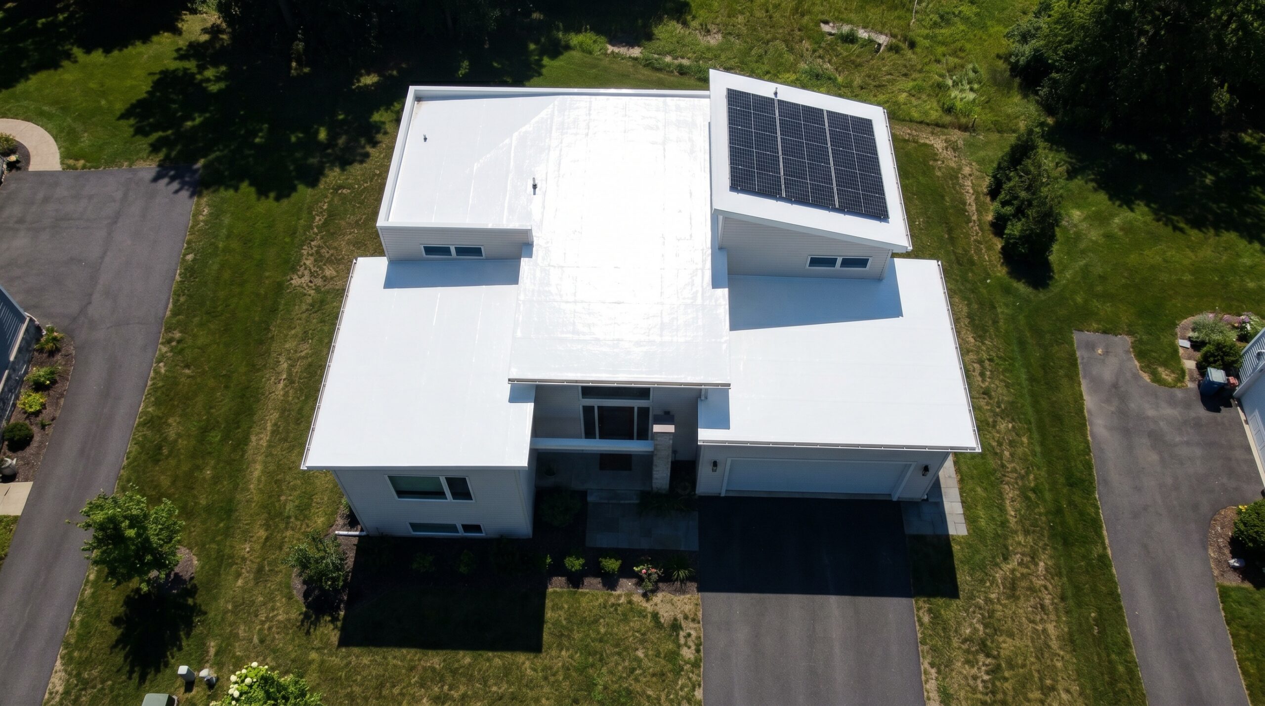 Aerial view of a modern white flat-roofed house with solar panels on one section of the roof, surrounded by grass lawns and driveways on either side.