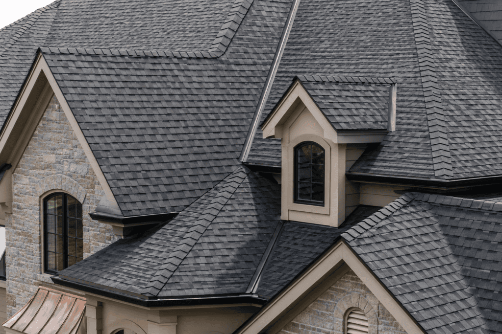 Close-up of a complex, multi-level roof with gray asphalt shingles, dormer windows, and beige trim on a stone house.