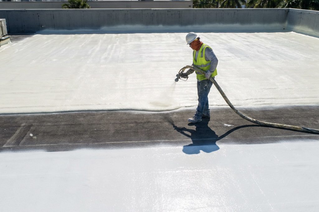 A construction worker in a hard hat and safety vest sprays white coating on a flat rooftop, covering dark roofing material with a light protective layer.