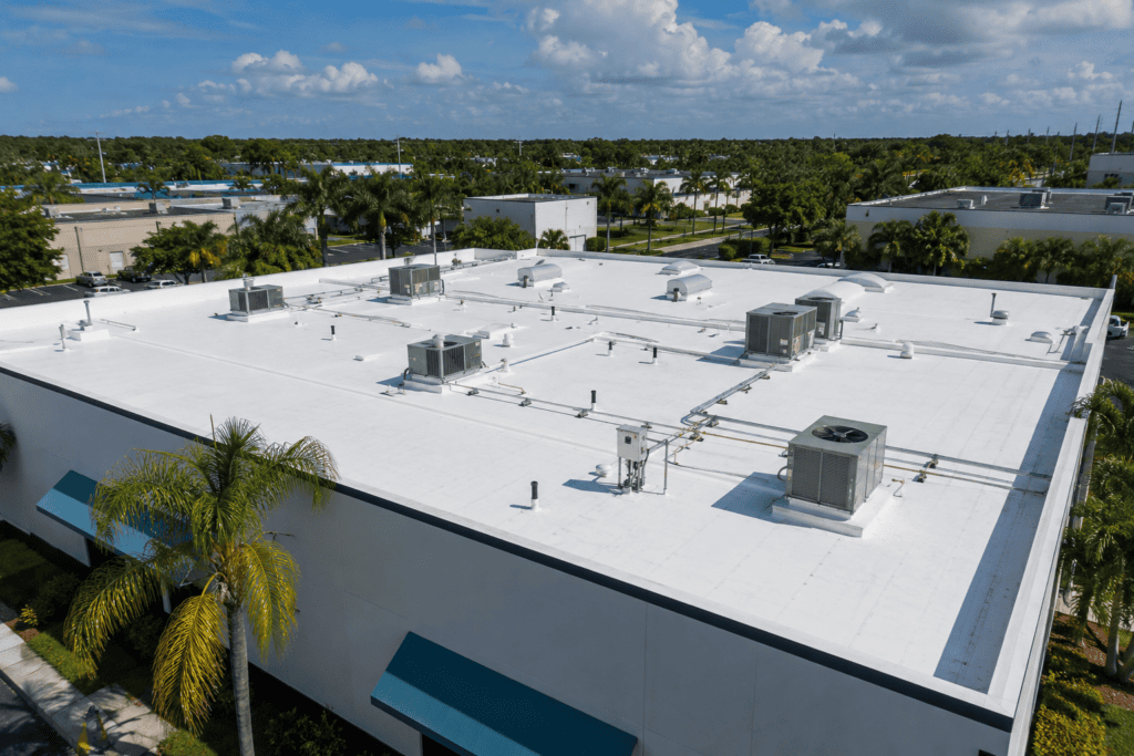Aerial view of a white flat-roofed commercial building with multiple rooftop HVAC units, surrounded by palm trees and other buildings under a partly cloudy sky.
