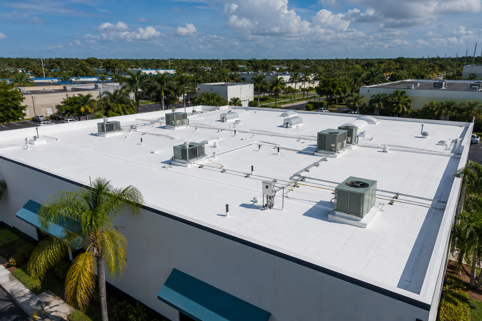 Aerial view of a white flat-roofed commercial building with multiple rooftop HVAC units, surrounded by palm trees and other buildings under a partly cloudy sky.