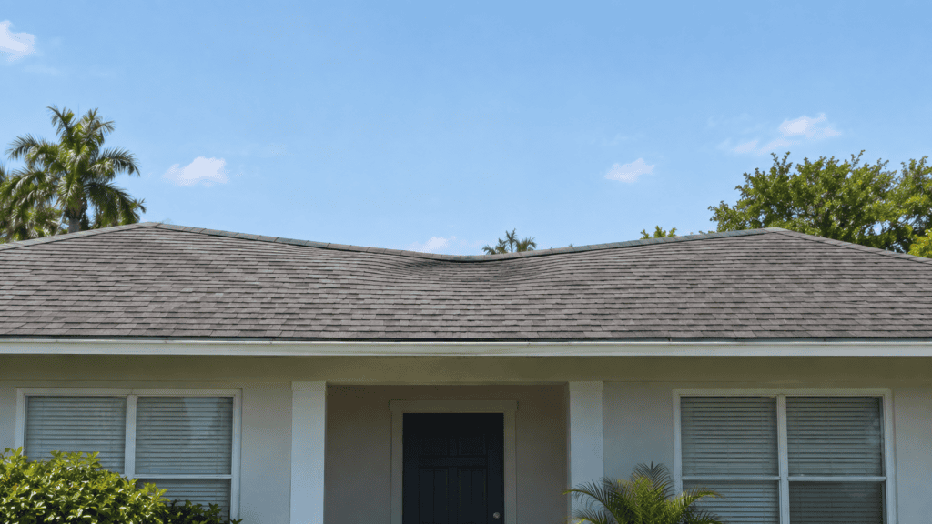 A house with a dark shingle roof showing a noticeable sag in the middle, white walls, two sets of closed white blinds, green bushes, palm trees, and a clear blue sky in the background.