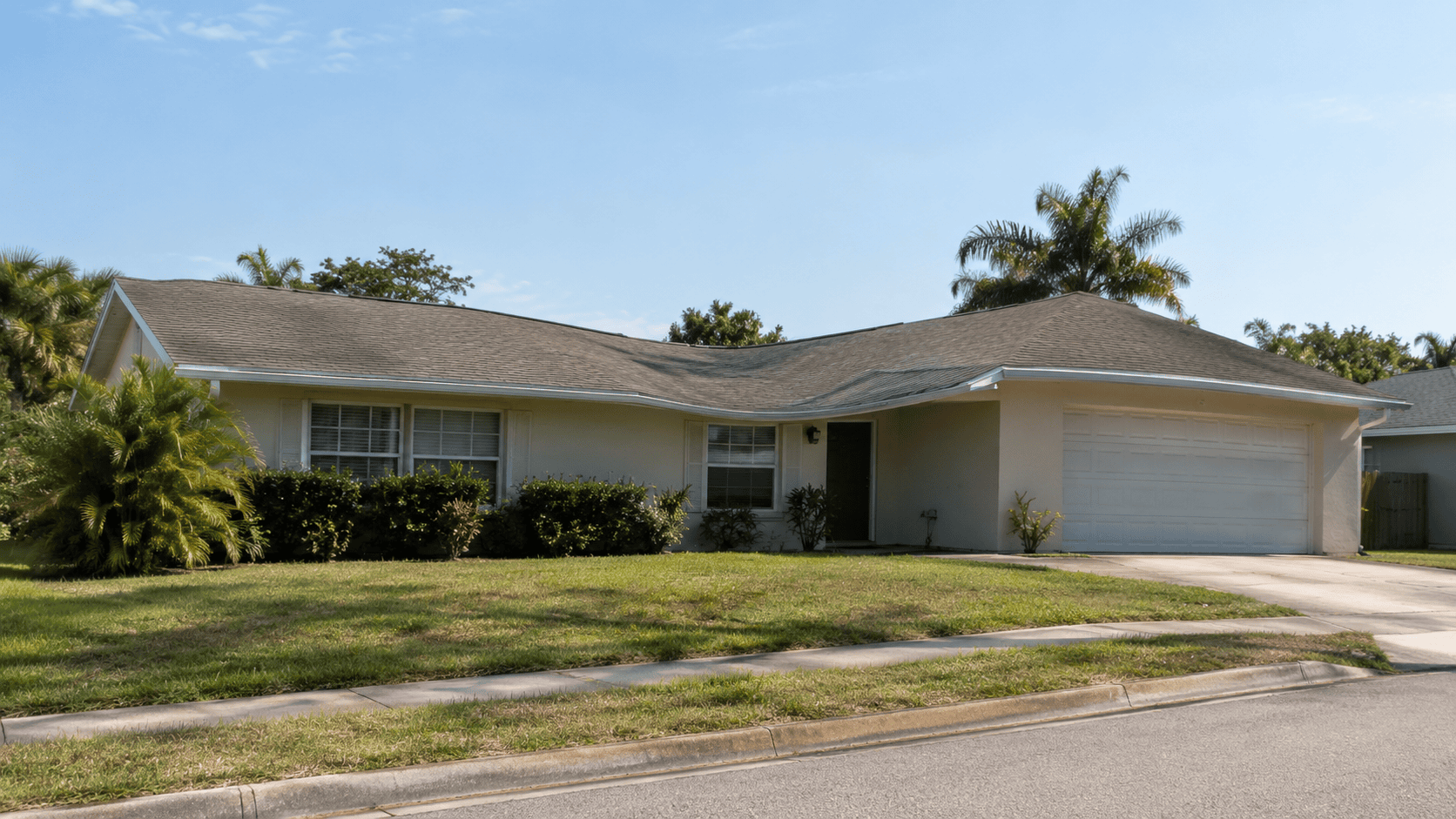 Single-story beige house with a gray roof, two front windows, bushes along the facade, a double garage, and a driveway. Palm trees and grass yard surround the house under a clear sky.